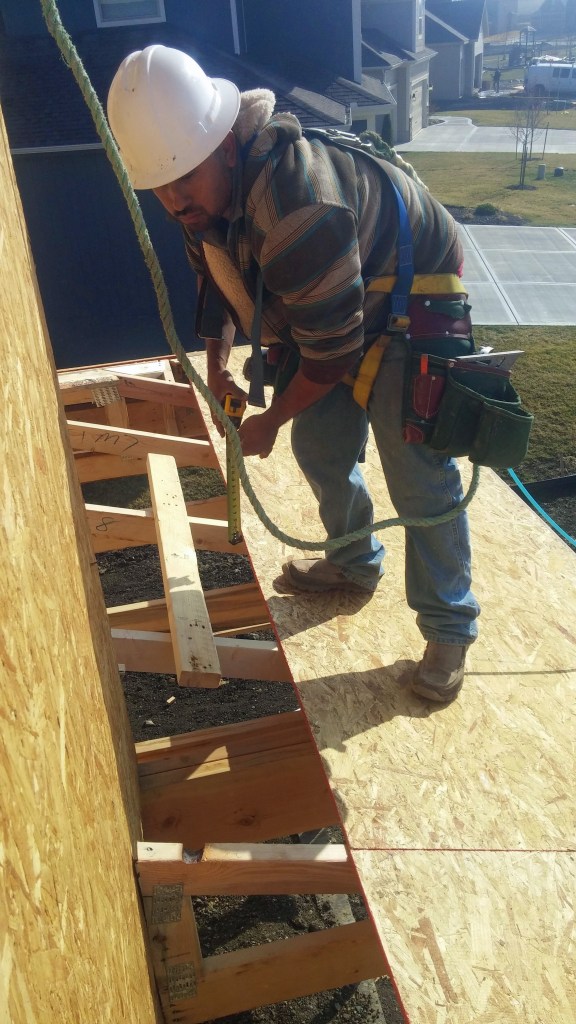On a Summit Custom Homes jobsite, a framing carpenter wears a safety harness secured to a point above him while working on the roof. He is wearing a hardhat and shoes that have good traction for the slope of the
roof. &nbsp;