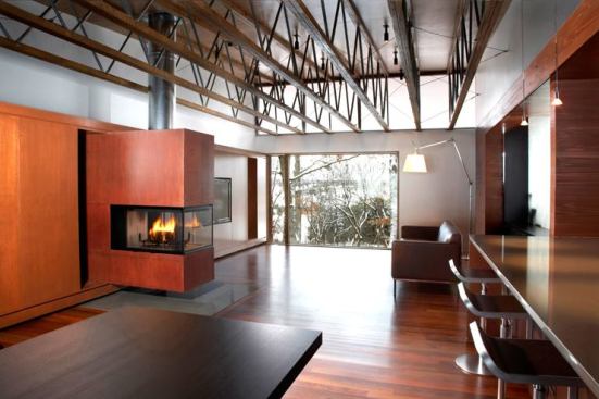 The house's new shed roof, raised above the structure's original roofline, is supported by exposed metal and wood trusses. The renovated interiors are a series of sleek, spacious, interconnected living spaces finished with warm, modern materials.
