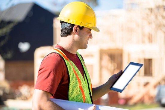 A Latin descent construction worker, supervisor, contractor, inspector, architect, or engineer inspects the work at a construction site.  Framed house, building in background.  He is wearing a hard hat and safety vest and holds a digital tablet and blueprints.
